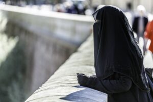 A woman in black abaya and veil wearing hand gloves standing by the wall.