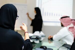 A woman teaching on the board while a woman and man are sitting.