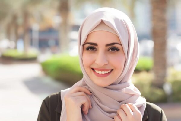 A woman in hijab is smiling. There is hedge of plants and trees behind her.